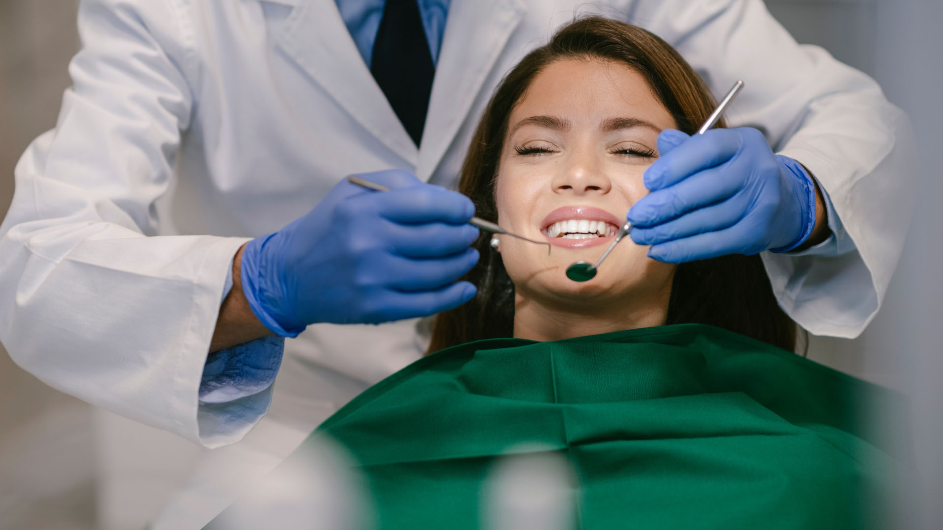 A dentist in blue gloves using dental instruments to examine a smiling woman wearing a green bib in a dental chair.